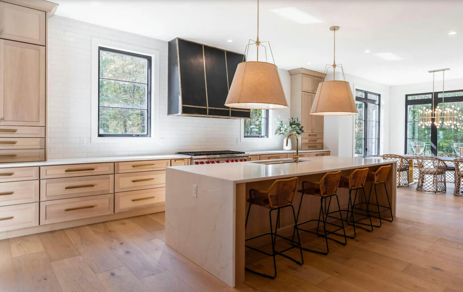 A modern farmhouse kitchen with light stained white oak cabinets, brass accents, black window frames and metal hood. The kitchen island has waterfall countertops and large scale fabric pendant lights.