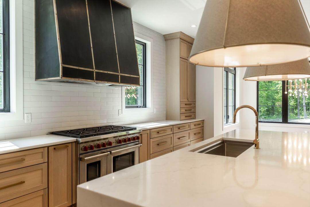 A shaker style kitchen with light stained wood cabinets using Quarter-Sawn White Oak wood. The interior design is accented with brass, black, and soft metal accents and the light fixtures use a natural linen like fabric with a modern farmhouse style.
