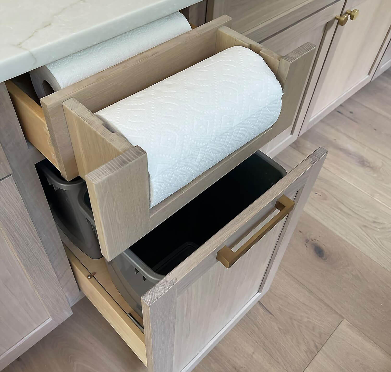 White Oak cabinets shown close up with a pull-out trash bin cabinet below a paper towel dispenser drawer that includes additional drawer storage for extra paper towels and misc. kitchen storage.