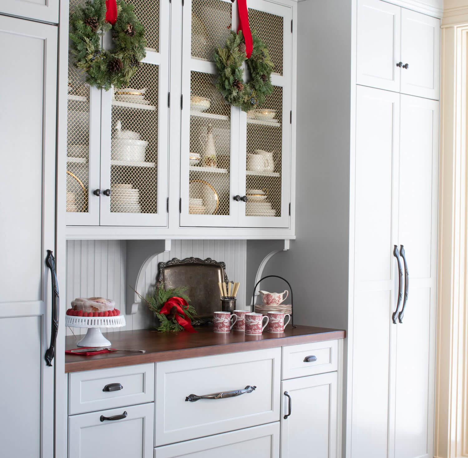 A beautiful butler's pantry space with the kitchen fridge hidden behind appliance panels. The wire mesh cabinet doors are decorated for the holidays with small wreaths.