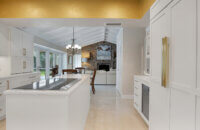 This bright white kitchen features Dura Supreme Cabinetry’s Homestead Panel door style in “White” paint paired with white countertops & backsplash with a golden ceiling and gold hardware pulls.
