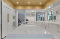 This bright white kitchen features Dura Supreme Cabinetry’s Homestead Panel door style in “White” paint paired with white countertops & backsplash with a golden ceiling and gold hardware pulls.