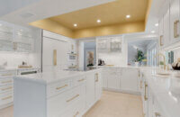 This bright white kitchen features Dura Supreme Cabinetry’s Homestead Panel door style in “White” paint paired with white countertops & backsplash with a golden ceiling and gold hardware pulls.