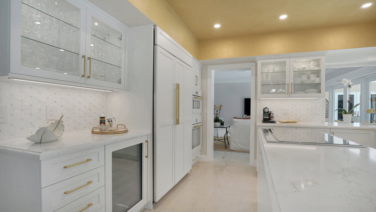 This bright white kitchen features Dura Supreme Cabinetry’s Homestead Panel door style in “White” paint paired with white countertops & backsplash with a golden ceiling and gold hardware pulls.