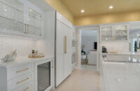 This bright white kitchen features Dura Supreme Cabinetry’s Homestead Panel door style in “White” paint paired with white countertops & backsplash with a golden ceiling and gold hardware pulls.