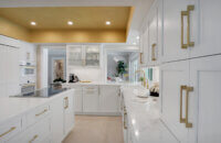 This bright white kitchen features Dura Supreme Cabinetry’s Homestead Panel door style in “White” paint paired with white countertops & backsplash with a golden ceiling and gold hardware pulls.