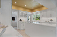This bright white kitchen features Dura Supreme Cabinetry’s Homestead Panel door style in “White” paint paired with white countertops & backsplash with a golden ceiling and gold hardware pulls.