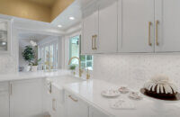 This bright white kitchen features Dura Supreme Cabinetry’s Homestead Panel door style in “White” paint paired with white countertops & backsplash with a golden ceiling and gold hardware pulls.