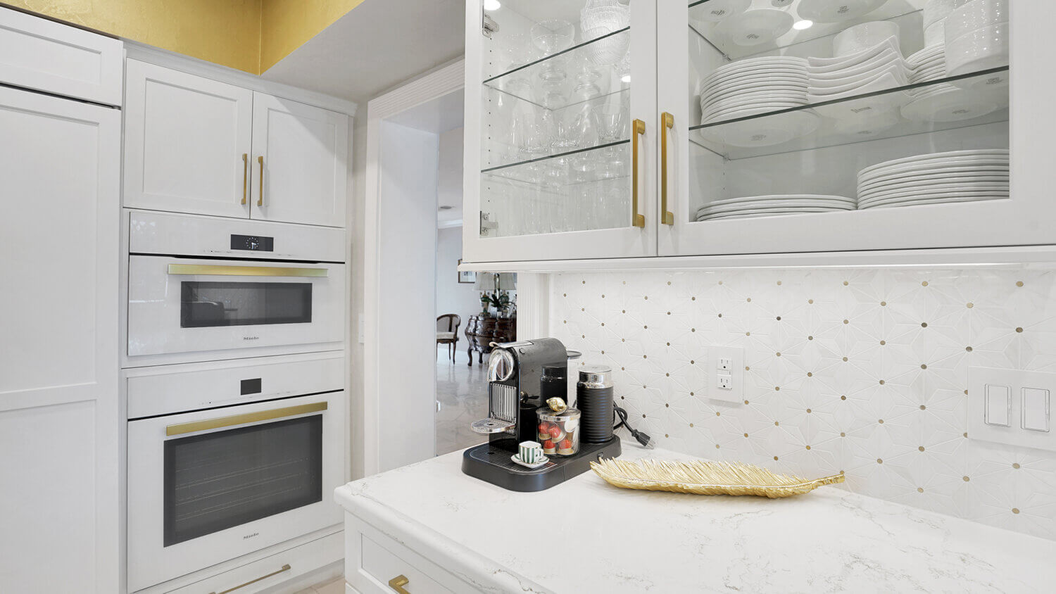 This bright white kitchen features Dura Supreme Cabinetry’s Homestead Panel door style in “White” paint paired with white countertops & backsplash with a golden ceiling and gold hardware pulls.