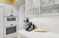 This bright white kitchen features Dura Supreme Cabinetry’s Homestead Panel door style in “White” paint paired with white countertops & backsplash with a golden ceiling and gold hardware pulls.