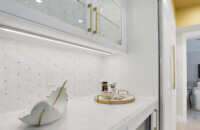 This bright white kitchen features Dura Supreme Cabinetry’s Homestead Panel door style in “White” paint paired with white countertops & backsplash with a golden ceiling and gold hardware pulls.
