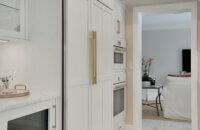This bright white kitchen features Dura Supreme Cabinetry’s Homestead Panel door style in “White” paint paired with white countertops & backsplash with a golden ceiling and gold hardware pulls.