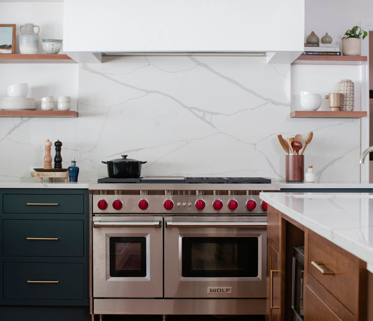 This kitchen is a great example of the latest trend of deep, earthy green cabinetry. Green is the color of nature, and it symbolizes growth, harmony, freshness, and fertility. This kitchen showcases Dura Supreme Cabinetry with the Homestead Panel Inset door style with optional solid slab drawer fronts in a combination of the “Rock Bottom” painted finish (perimeter cabinets) and the “Toast” stain on Cherry (kitchen island & floating shelves).