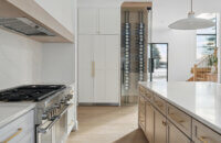 A luxurious modern farmhouse design with a range alcove, a light wood kitchen island, and a wine bottle wall behind glass.