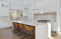 A stunning modern farmhouse kitchen with a wide range alcove space with a simple raw-wood panel mantel over the range. The island features custom fluted details to accent the seating area while waterfall countertops frame the island.