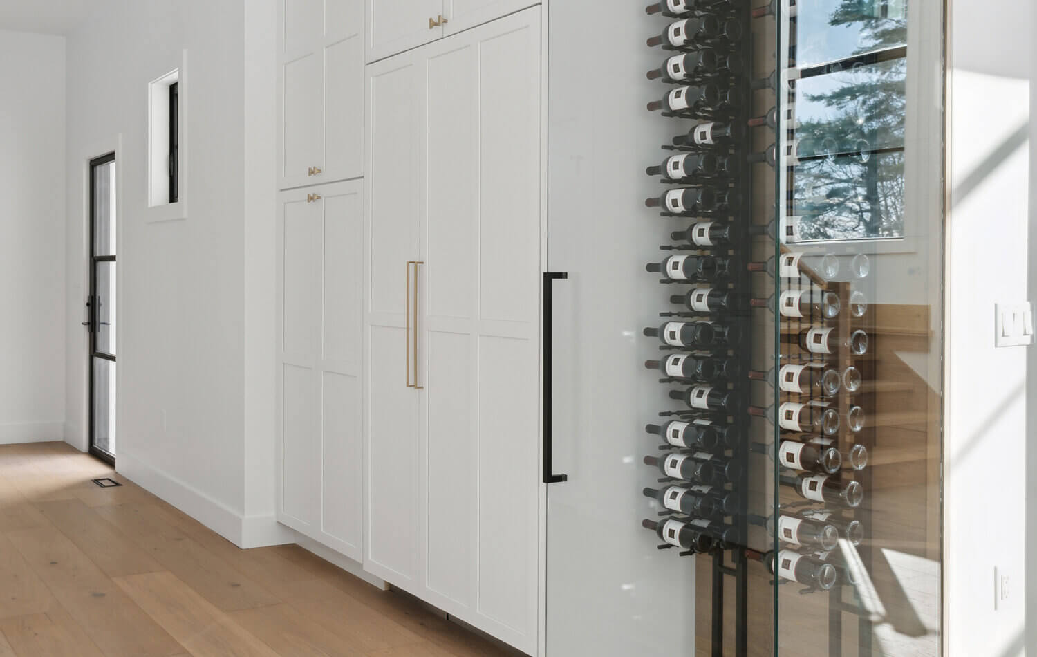 A floor-to-ceiling wine rack wall behind glass. The wine storage is accented with light stained quarter-sawn white oak wood that beautifully contrasts the white painted cabinets throughout the kitchen. The large refrigerator is hidden behind cabinet door appliance panels.