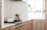 A modern farmhouse kitchen featuring light stained quarter-sawn white oak wood cabinets with a sleek micro, skinny shaker doors style and bright white countertops. A white range alcove frames the cooking area.