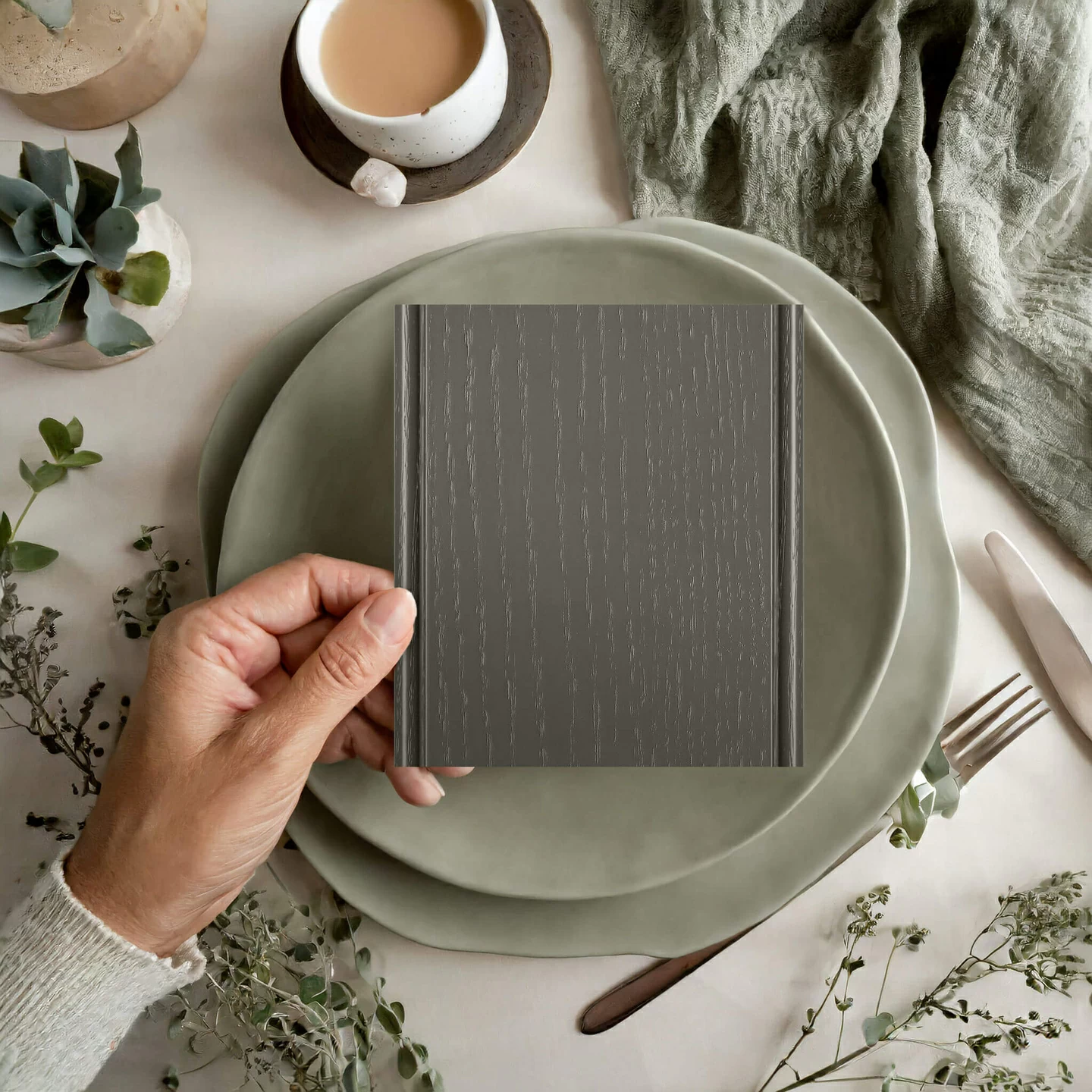 A kitchen design mood board with a hand holding a sample of painted oak in a earthy green painted finish with a defined wood grain texture.